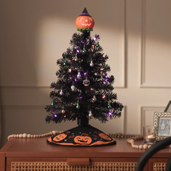 A black miniature holiday tree decorated with mini Halloween ornaments and a light-up jack-o-lantern tree topper that plays music sits on a wooden side table.